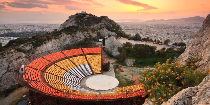 Lycabettus Theatre at sunset, colourful open-air amphitheatre carved into the hillside with Athens and the chapel visible above